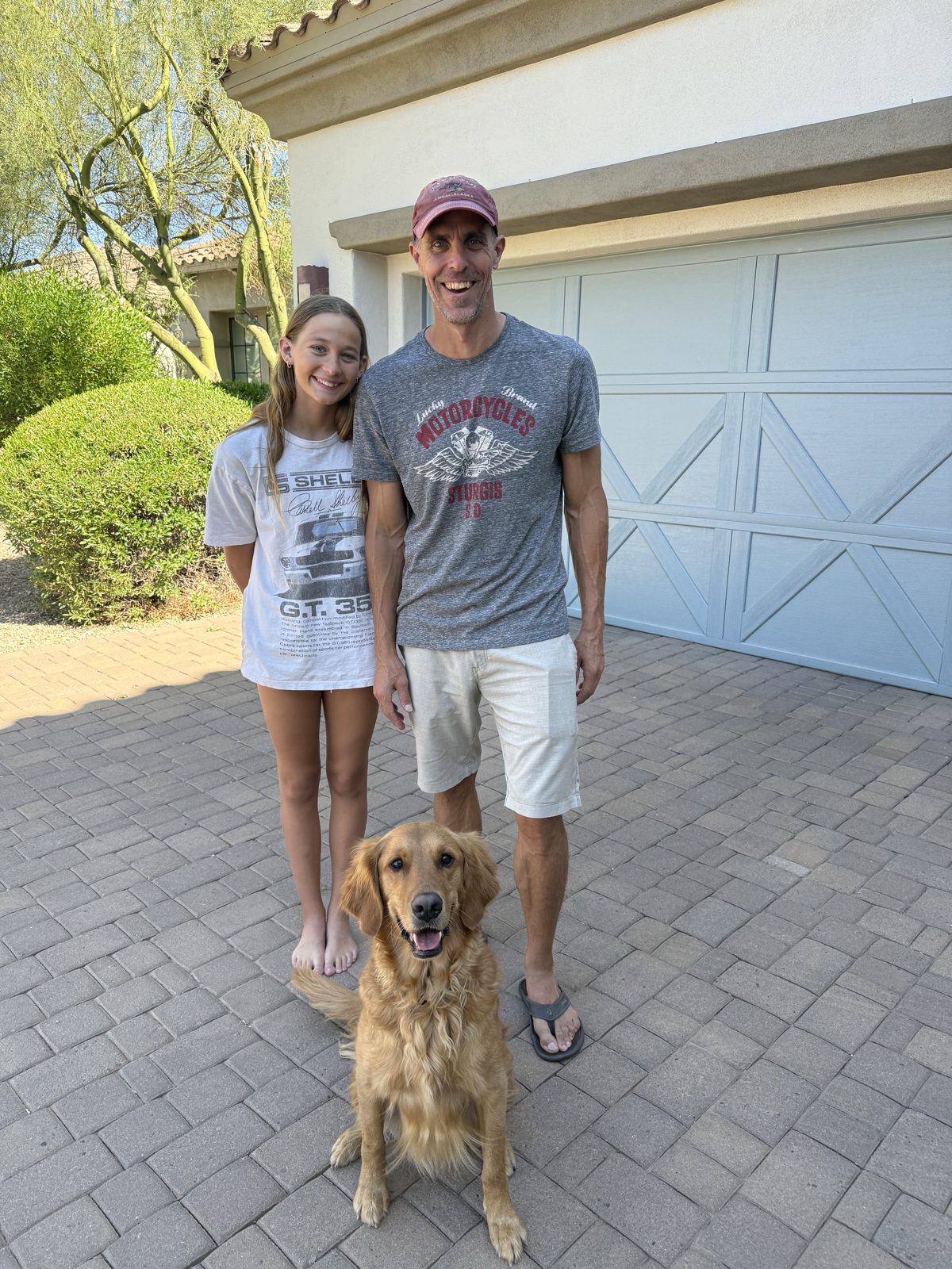 Golden retriever sitting with eyes locked on the camera between two smiling family members on a sunny driveway