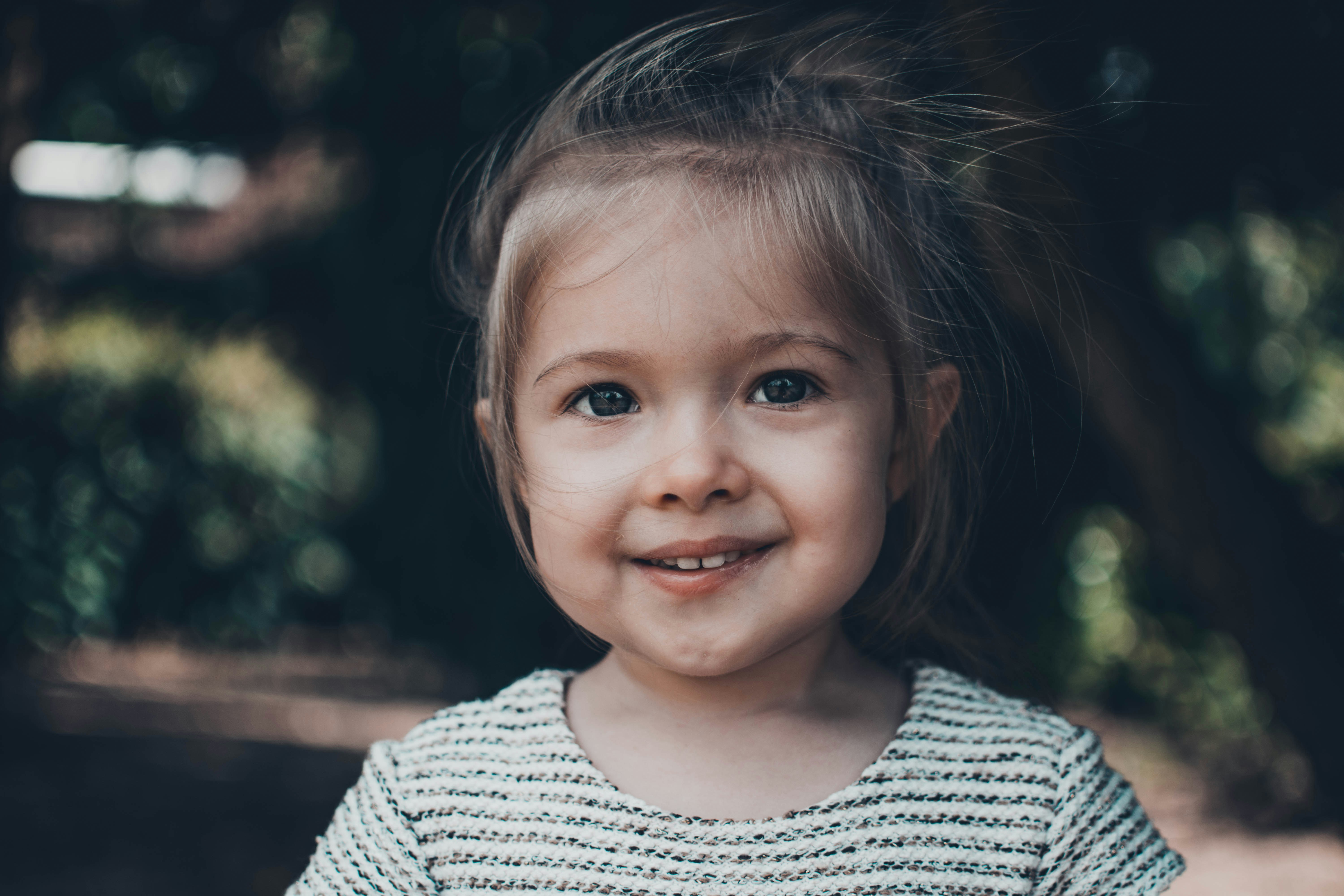 A smiling two-year-old girl outdoors in soft natural light, looking toward the camera with a real grin